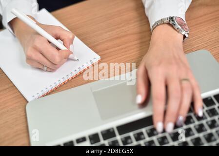 Primo piano delle mani femminili che scrivono in blocco note e che scrivono sopra tastiera per computer portatile Foto Stock