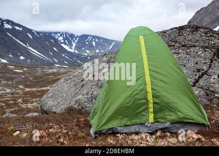 Verde impermeabile e vento tenda per escursioni è in montagna a tempo ventoso, in piedi dietro grande pietra per la protezione dal vento, copia spazio Foto Stock