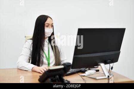Una giovane dipendente della Banca in una maschera facciale funziona con un computer desktop in ufficio Foto Stock