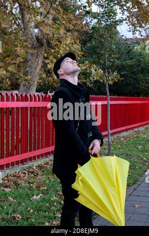 Uomo in camice nero, in piedi per le strade della città con ombrello giallo tra le mani, guardando il cielo per gocce di pioggia. Previsioni e tempo piovoso in autunno. Foto Stock