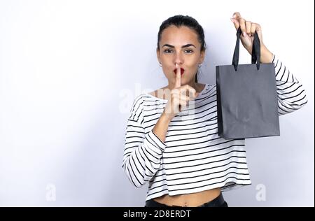 Giovane bella donna che tiene una borsa nera della spesa chiedendo di essere tranquilla con il dito sulle labbra. Silenzio e concetto segreto. Black Friday Foto Stock