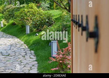 Vista prospettica attraverso il cancello di legno aperto sfocato con decorazioni in metallo A passerella in pietra nel giardino giapponese Zen fiancheggiato da ornamentali piante e lanterne Foto Stock