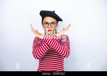 Giovane bella donna bruna che indossa beretto francese e occhiali su sfondo bianco espressione di rifiuto che attraversa le braccia facendo segno negativo, fa arrabbiato Foto Stock