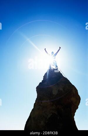 Un uomo che festeggia con le mani sulla testa in cima a un ripido pinnacolo di roccia con il sole che si alza da dietro. Foto Stock