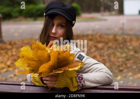 Primo piano ritratto di una giovane donna dai capelli castani, con cappuccio nero e cappotto bianco. Sullo sfondo di un parco autunnale. Con foglie gialle Foto Stock