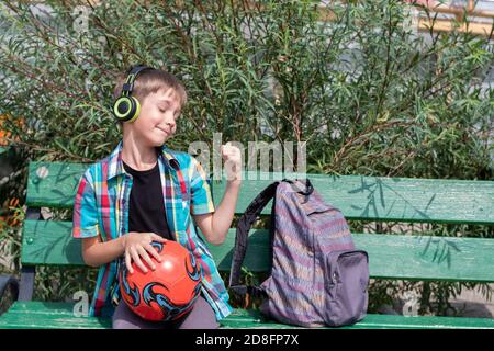 Un simpatico ragazzo di scuola ascolta la musica con le cuffie, si siede su una panchina, accanto a una palla e una borsa Foto Stock