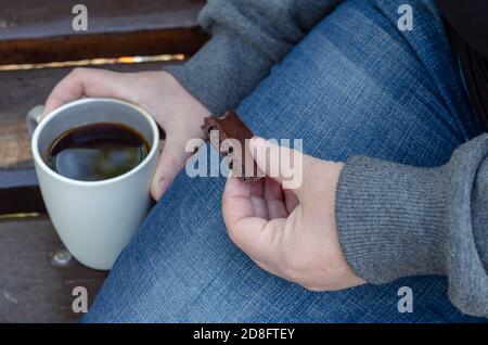 Una donna adulta beve caffè e mangia bar di cioccolato su panca. Tazza di caffè in ceramica e pezzo di cioccolato nero in mano. Vista dall'alto Foto Stock