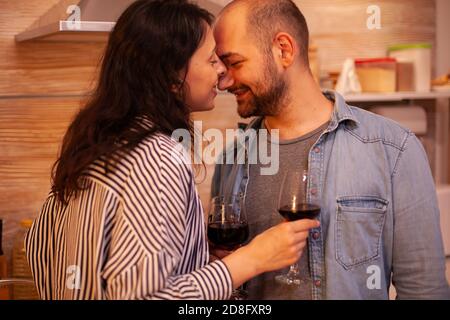 Coppia che tiene gli occhi chiusi durante la data romantica in cucina. Coppia adulta a casa, bere vino rosso, parlare, sorridere, gustare il pasto in sala da pranzo. Foto Stock