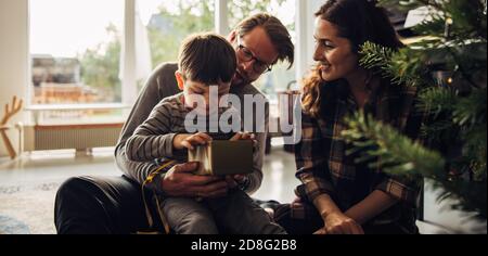 Bambino seduto con i genitori che aprono la scatola del regalo mentre si siede vicino all'albero di natale. La giovane famiglia festeggia il natale con i regali. Foto Stock