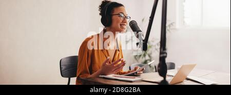 Donna che registra un podcast sul suo computer portatile con cuffie e un microscopio. Podcaster femminile che crea podcast audio dal suo studio di casa. Foto Stock