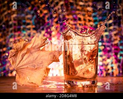 cubetti di ghiaccio che spruzzano all'interno del bicchiere di whisky con la foglia di acero a. sfondo colorato Foto Stock