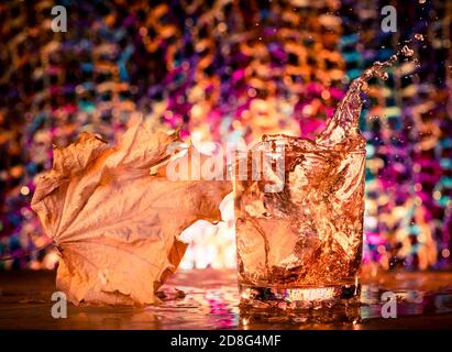 cubetti di ghiaccio che spruzzano all'interno del bicchiere di whisky con la foglia di acero a. sfondo colorato Foto Stock