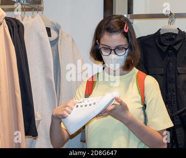 Bella ragazza caucasica in una maschera sceglie scarpe e vestiti in un negozio. Il concetto di shopping durante una pandemia Foto Stock