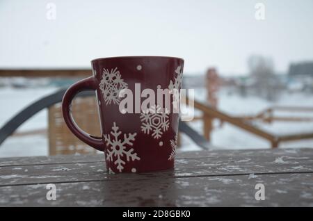 Coppa rossa o borgogna con un modello di fiocchi di neve sullo sfondo di una foresta invernale. Nevicare. Capodanno. Natale. Tazza con una bevanda calda sul Foto Stock