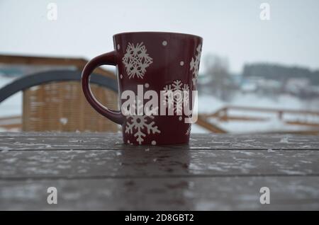 Coppa rossa o borgogna con un modello di fiocchi di neve sullo sfondo di una foresta invernale. Nevicare. Capodanno. Natale. Tazza con una bevanda calda sul Foto Stock