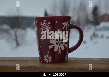 Coppa rossa o borgogna con un modello di fiocchi di neve sullo sfondo di una foresta invernale. Nevicare. Capodanno. Natale. Tazza con una bevanda calda sul Foto Stock