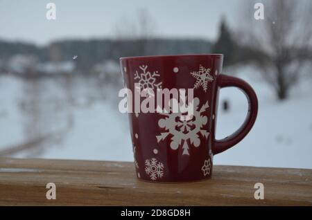 Coppa rossa o borgogna con un modello di fiocchi di neve sullo sfondo di una foresta invernale. Nevicare. Capodanno. Natale. Tazza con una bevanda calda sul Foto Stock