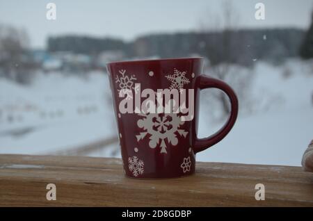 Coppa rossa o borgogna con un modello di fiocchi di neve sullo sfondo di una foresta invernale. Nevicare. Capodanno. Natale. Tazza con una bevanda calda sul Foto Stock