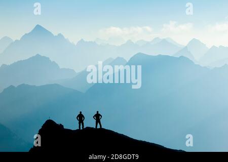 Due uomini che raggiungono la vetta dopo l'arrampicata e le escursioni godendo la libertà e guardando verso le montagne sagome panorama durante l'alba. Foto Stock