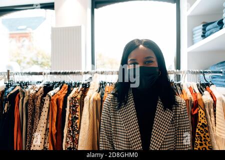 ritratto di una bella donna nera che indossa una maschera facciale nel suo negozio di moda. Un'allegra lavoratrice che si trova di fronte alla fotocamera in boutique Foto Stock