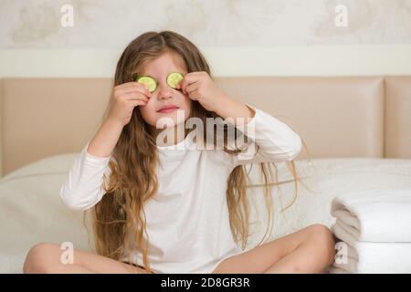 Ritratto di una ragazza sorridente felice. Cute 5 anni ragazza che si rilassa sul letto e facendo maschera facciale con fette di cetriolo. Concetto di cura della pelle. Foto Stock