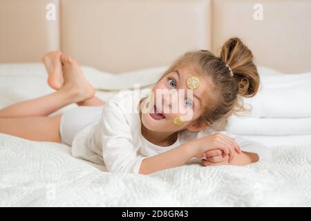 Ritratto di una ragazza sorridente felice. Cute 5 anni ragazza che si rilassa sul letto e facendo maschera facciale con fette di cetriolo. Concetto di cura della pelle. Foto Stock