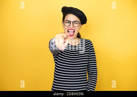 Giovane bella donna bruna che indossa beretto francese e occhiali su sfondo giallo ridendo a voi, puntando il dito alla macchina fotografica con la mano sopra b Foto Stock