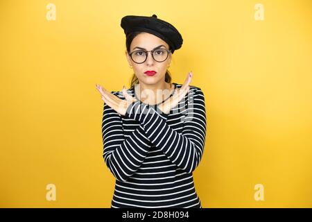Giovane bella donna bruna indossando beretto francese e occhiali su sfondo giallo espressione reiezione che attraversa le braccia facendo segno negativo, arrabbiato f Foto Stock