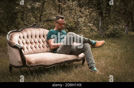 Uomo riposante in jeans grigi seduto su un divano dentro il mezzo del giardino Foto Stock
