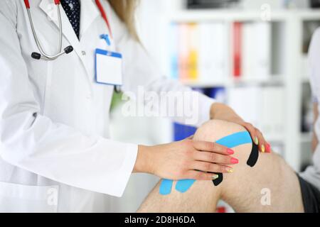 Il Traumatologo si incolla con nastro blu e nero sul ginocchio irritato del paziente in primo piano della clinica Foto Stock