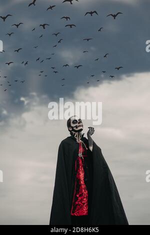 La giovane donna si trova in costume di morte di Halloween con scheletro dipinto sul corpo contro il cielo scuro e i corvi volanti. Concetto di Halloween. Cranio di zucchero Foto Stock