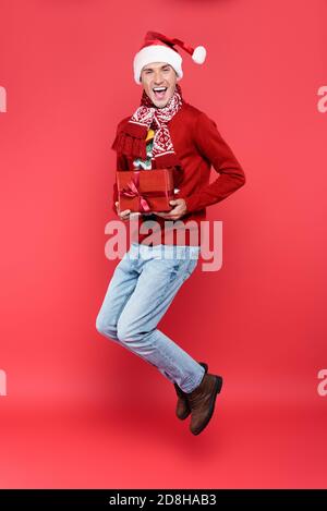 Uomo allegro nel cappello di santa che tiene la scatola del regalo mentre salta su sfondo rosso Foto Stock