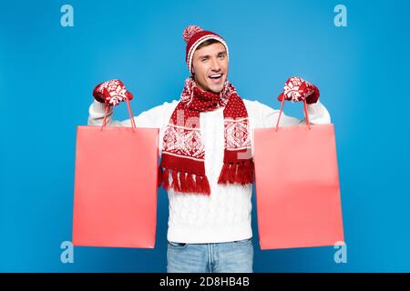 Felice giovane uomo adulto in mittens che tiene sacchetti di carta rossi isolato su blu Foto Stock