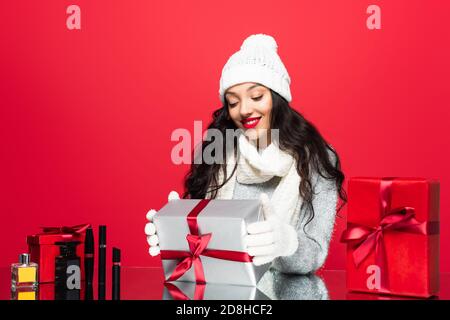 gioiosa donna in cappello, guanti e sciarpa calda che tiene regalo di natale vicino cosmetici decorativi isolati sul rosso Foto Stock