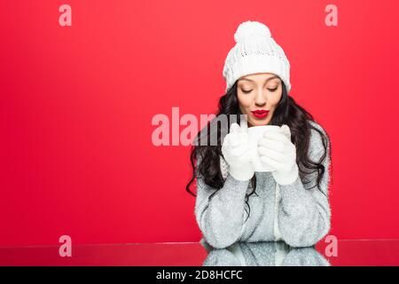 donna felice in outfit invernale soffiando sulla tazza con bevanda isolato in rosso Foto Stock