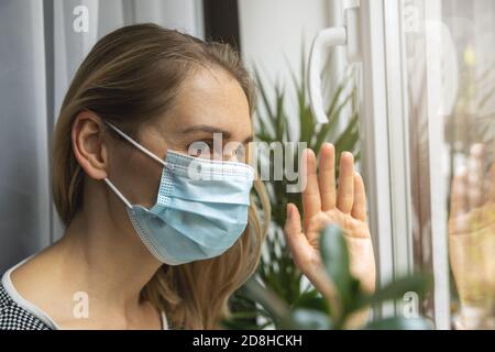 home quarantena virus pandemic - giovane donna con viso medico maschera guardando attraverso la finestra e ondeggiando con la mano ad un amico Foto Stock