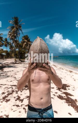 Un uomo tiene un cocco fresco di fronte alla sua testa su una spiaggia tropicale vuota a Marie Galante, Guadalupa Foto Stock