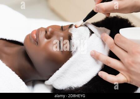 Vista ravvicinata dell'uomo che applica la maschera facciale con il cosmetico spazzola sulla fronte di donna afroamericana nel salone termale Foto Stock
