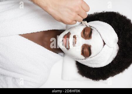 vista dall'alto della donna afro-americana positiva che indossa accappatoio, sdraiata vicino al terapeuta termale applicando la maschera sulla guancia nel salone termale Foto Stock