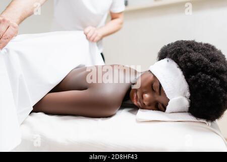 Massaggiatore che copre donna afroamericana con lenzuola in salone termale, striscione Foto Stock