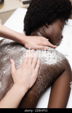Primo piano vista del massaggiatore che massaggiava la donna afroamericana con scrub nel salone termale Foto Stock
