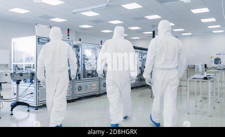 Back Shot di tre scienziati in Protective Coverall's Walking in sterile High Precision Manufacturing Laboratory per accendere Machninery, utilizzare Foto Stock