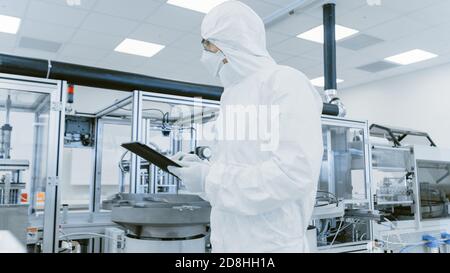 Controllo di qualità: Lo scienziato che utilizza il computer Digital Tablet e indossa un tuta protettiva attraversa il laboratorio di produzione. Prodotto Foto Stock
