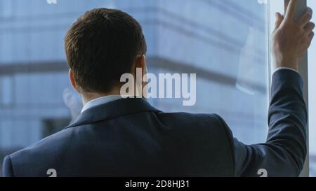 Primo piano di un uomo d'affari pensieroso in una tuta in piedi nel suo ufficio e contemplare il prossimo grande affare di affari, guardando fuori dalla finestra. Grande Foto Stock