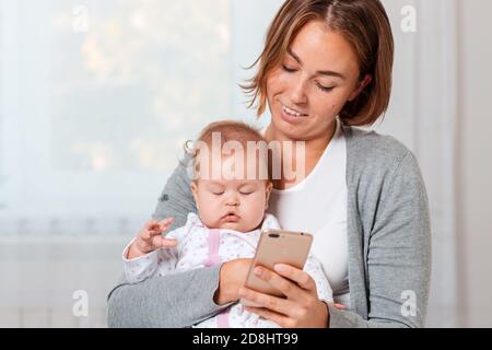 Una madre tiene un bambino addormentato tra le braccia e gli mostra qualcosa sul suo smartphone. Primo piano verticale. Foto Stock