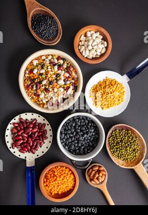 Legumi vari impulsi su sfondo nero del piano portapaziente. Scoop, ciotole e mestoli con cibo sano vegetariano secco. Vista dall'alto, disposizione piatta Foto Stock