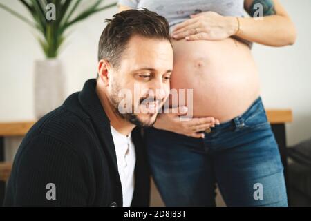 Felice giovane uomo che mette l'orecchio al ventre delle donne incinte ascoltando il bambino che si muove all'interno. Foto Stock