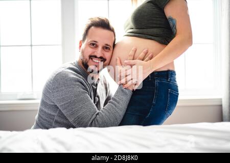 Felice giovane uomo che mette l'orecchio al ventre delle donne incinte ascoltando il bambino che si muove all'interno. Foto Stock