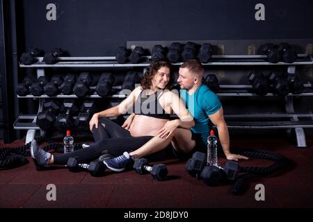 Donna incinta va per gli sport con suo marito in palestra per cross training. Coppia atleti che si aspettano il bambino mentre riposano dopo l'addestramento e gli esercizi Foto Stock