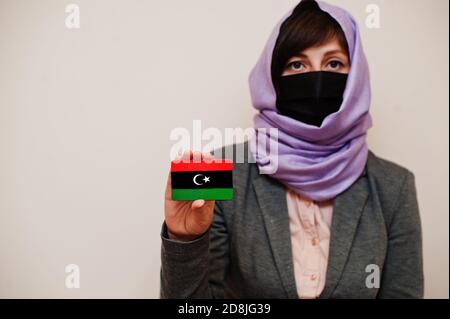 Ritratto di giovane donna musulmana che indossa abiti formali, proteggi maschera facciale e sciarpa testa hijab, tieni la carta bandiera Libia su sfondo isolato. Coronavi Foto Stock